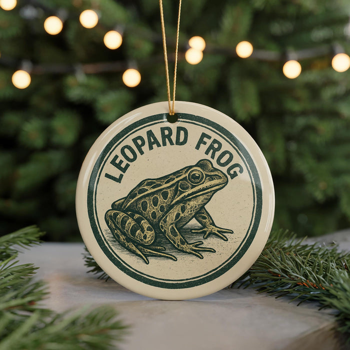 A round ornament featuring a leopard frog design, hanging from a string against a blurred background of a Christmas tree with lights.