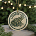 A round ornament featuring a leopard frog design, hanging from a string against a blurred background of a Christmas tree with lights.