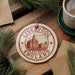 A circular ornament with the text "LIVERPOOL ENGLAND" and an illustration of a building, placed on a wooden surface next to a cup of coffee and some pine branches.