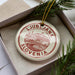 A circular ornament with the text "LJUBBLJANA SLOVENIA" printed on it, placed inside a box with pine branches.