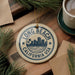 A circular ornament with the text "LONG BEACH CALIFORNIA USA" printed on it, surrounded by pine branches and a cup of coffee on a wooden surface.