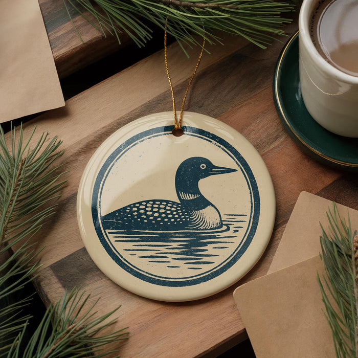 a ceramic ornament with a blue and white design depicting a loon, which is a type of bird, sitting on a lake. The ornament is hanging from a string and is placed on a wooden surface, surrounded by pine branches and a cup of coffee.