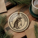 A decorative ornament featuring a penguin is displayed on a wooden surface, surrounded by pine branches and a cup of coffee.
