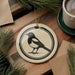 a round ceramic ornament with a black and white illustration of a bird, likely a magpie, on it. The ornament is placed on a wooden surface, surrounded by pine branches and a cup of coffee.
