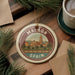 A circular ornament with the text "MALAGA SPAIN" and an illustration of a cityscape, surrounded by pine branches and a cup of coffee on a wooden surface.