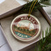 A circular ornament with the text "MALAGA SPAIN" printed on it, surrounded by palm trees and a building, is displayed in a box with pine branches.