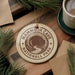 A round ornament with the text "MAMMOTH CAVE NATIONAL PARK" printed on it, surrounded by pine branches and a cup of coffee on a wooden surface.