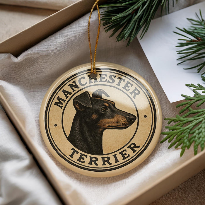 A round ornament featuring a black and tan dog's head, with the text "MANCHESTER TERRIER" printed on it, is displayed in a box with pine branches.