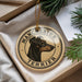 A round ornament featuring a black and tan dog's head, with the text "MANCHESTER TERRIER" printed on it, is displayed in a box with pine branches.