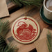 A round ornament featuring a red bird, possibly a duck, is displayed on a wooden surface alongside a cup of coffee and some papers.