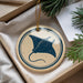 A round ornament with a blue stingray design is displayed in a box, surrounded by pine branches and a white card.