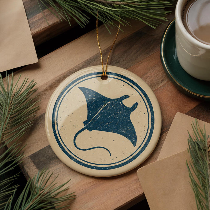 a round ornament with a blue stingray design, placed on a wooden surface next to a cup of coffee and some pine branches.