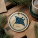 a round ornament with a blue stingray design, placed on a wooden surface next to a cup of coffee and some pine branches.