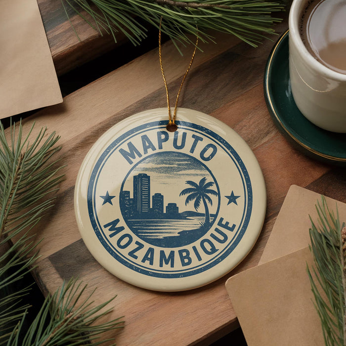 A circular ornament with the text "MOPUTO MOZAMBIQUE" and a graphic of a city skyline, hanging from a string. The ornament is placed on a wooden surface, surrounded by pine branches and a cup of coffee.