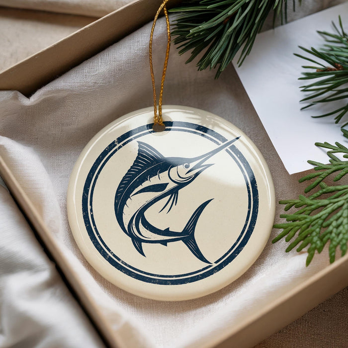 A round ornament with a blue and white design featuring a fish is displayed in a box, surrounded by pine branches.
