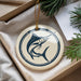 A round ornament with a blue and white design featuring a fish is displayed in a box, surrounded by pine branches.