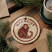 A round ornament featuring a monkey sitting on a branch, with the word "Marmoset" written on it. The ornament is placed on a wooden surface, surrounded by pine branches and a cup of coffee.
