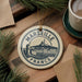A circular ornament with the text "MARSEILLE FRANCE" and an illustration of a cityscape is displayed on a wooden surface, surrounded by pine branches and a cup of coffee.
