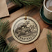 A circular ornament featuring a woodchuck illustration is displayed on a wooden surface, accompanied by a cup of coffee and pine branches.