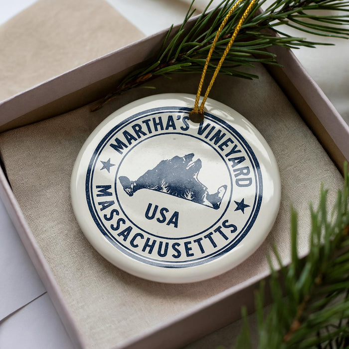 A white ornament with a blue border and text, featuring the name 'Martha's Vineyard' and the location 'Massachusetts USA', is displayed in a box with pine branches.