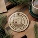 A circular ornament with the text "MEDINA SAUDI ARABIA" printed on it, surrounded by pine branches and a cup of coffee on a wooden surface.