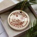 A circular ornament with the text "MERZOUGA MOROCCO" printed on it, placed in a box with pine branches.