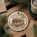 A circular ornament with the text "MESA VERDE NATIONAL PAARK USA" printed on it, surrounded by pine branches and a cup of coffee on a wooden surface.