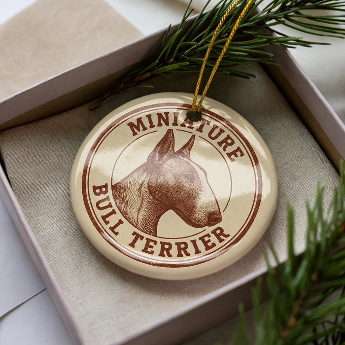 A circular ornament with a bull terrier image and the text "MINIATURE BULL TERRIER" printed on it, placed inside a box with pine branches.