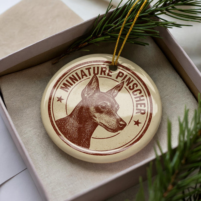 A round ornament with a dog's head and the text "MINIATURE PINSCHER" printed on it, placed inside a box with pine branches.