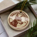 A round ornament with a dog's head and the text "MINIATURE PINSCHER" printed on it, placed inside a box with pine branches.