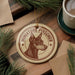 A round ornament featuring a miniature pinscher dog, with the text "MINIATURE PINSCHER" printed on it, is displayed on a wooden surface alongside a cup of coffee and some pine branches.