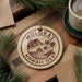 A circular ornament with the text "MOLOKA'I HAWAII USA" printed on it, surrounded by pine branches and a cup of coffee on a wooden surface.