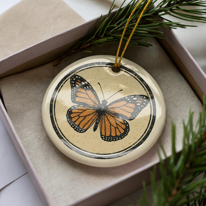 A ceramic ornament in the shape of a butterfly is displayed in a box, surrounded by pine branches.