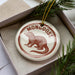A round ornament with a brown and white design featuring a moose, placed inside a box with pine branches.