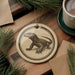 a round ornament with a lizard design, placed on a wooden surface next to a cup of coffee and some pine branches.
