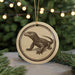 A circular ornament featuring a detailed illustration of a lizard or salamander, with a string hanging from it. The ornament is placed on a surface with pine branches in the background, creating a festive and nature-inspired setting.
