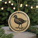 A round ornament featuring a black bird with a white stripe on its wing, sitting on a branch with pine needles.