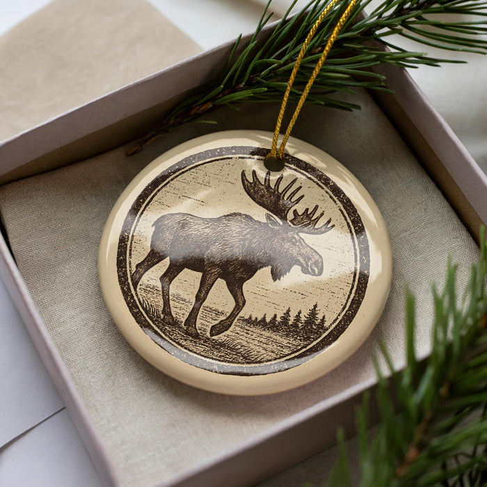 A circular ornament featuring a moose silhouette is displayed in a box, surrounded by pine branches.