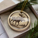 A circular ornament featuring a moose silhouette is displayed in a box, surrounded by pine branches.
