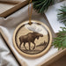 A circular ornament featuring a moose silhouette is displayed in a box, surrounded by pine branches.