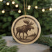 A circular ornament featuring a moose silhouette is displayed against a blurred background of Christmas lights.