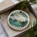 A circular ornament with a mountain landscape and a lake, surrounded by greenery, is displayed in a box.
