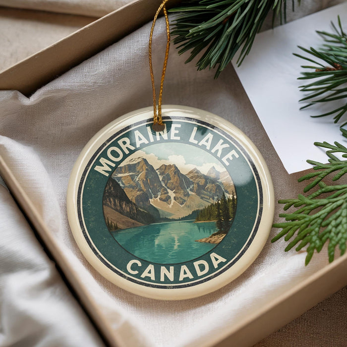 A round ornament with a mountain landscape and a lake, featuring the text "MORAIN LAKE CANADA" on it, is displayed in a box with pine branches.