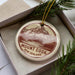 A round ornament with a mountain landscape design, placed in a box with pine branches.