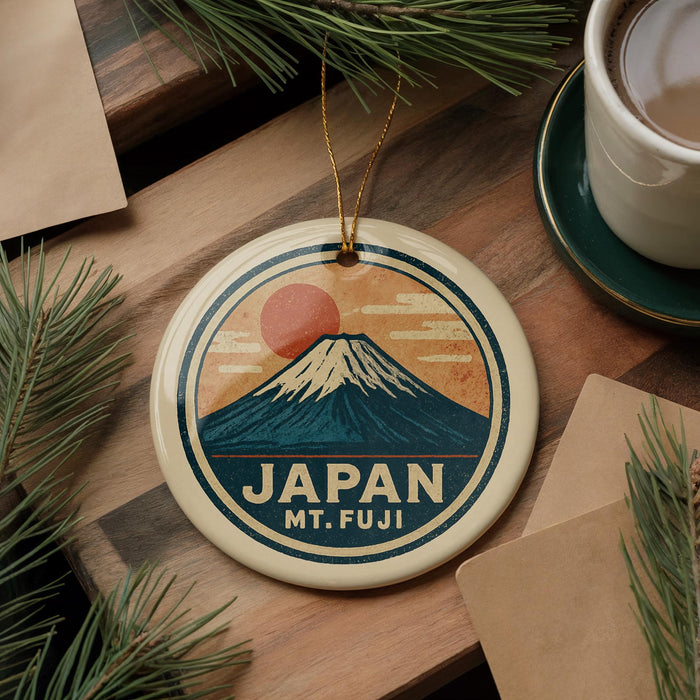A circular ornament featuring a mountain and sun design, with the text "JAPAN Mt. FUJI" printed on it, is displayed on a wooden surface alongside a cup of coffee and some pine branches.