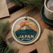 A circular ornament featuring a mountain and sun design, with the text "JAPAN Mt. FUJI" printed on it, is displayed on a wooden surface alongside a cup of coffee and some pine branches.