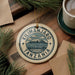 A decorative ornament with the text "KILIMANJARO TANZANIA" and an illustration of a mountain, placed on a wooden surface alongside a cup of coffee and some pine branches.