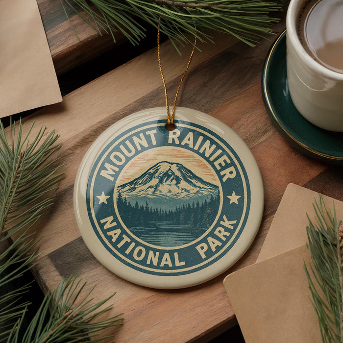 A circular ornament with the text "Mount Rainier National Park" and a mountain landscape design, placed on a wooden surface alongside pine branches and a cup of coffee.