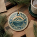 A circular ornament with the text "Mount Rainier National Park" and a mountain landscape design, placed on a wooden surface alongside pine branches and a cup of coffee.