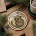A decorative ornament featuring a cartoon image of a mouse with the text "MOOSE LEMUR" on it, placed on a wooden surface alongside a cup of coffee and pine branches.